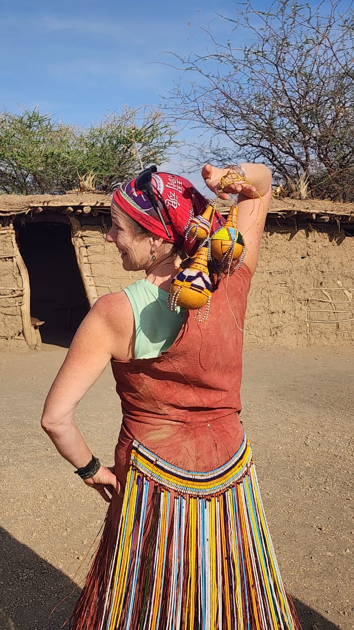 Guest in traditional Datoga dress holding beaded gourds outside a hut