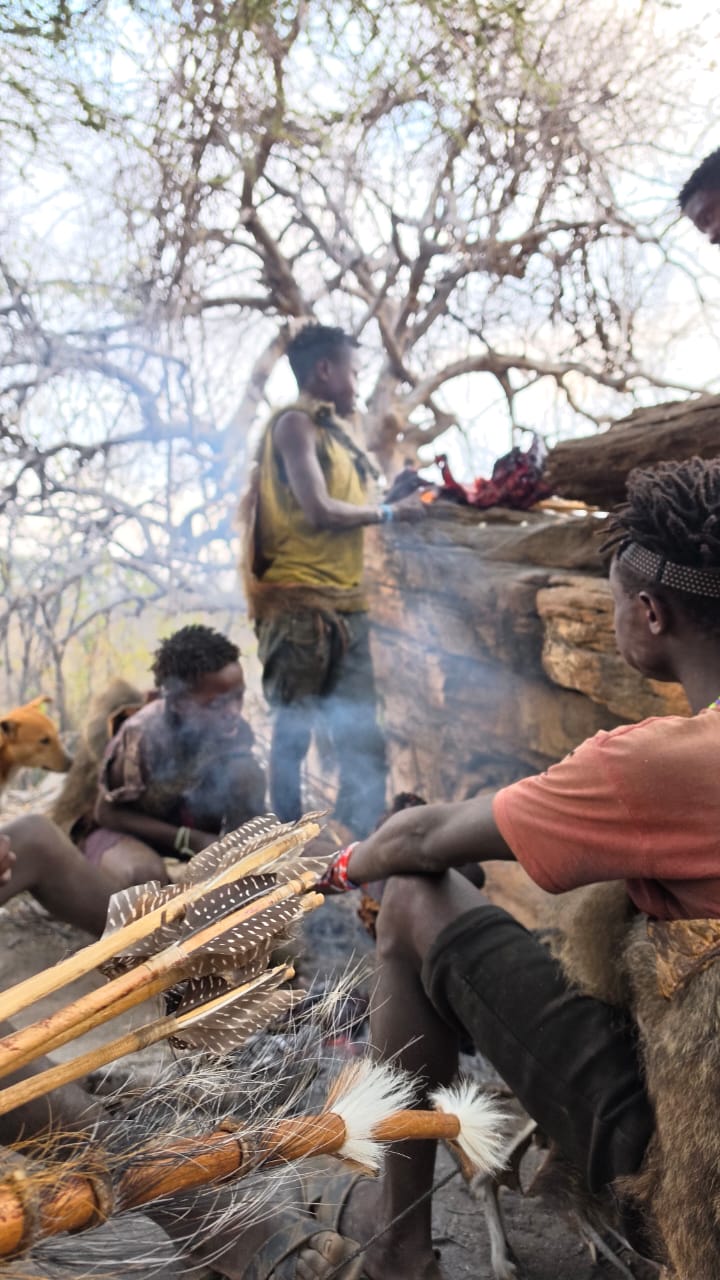 Feathered Hadzabe arrows in the foreground as hunters butcher the catch in wood smoke