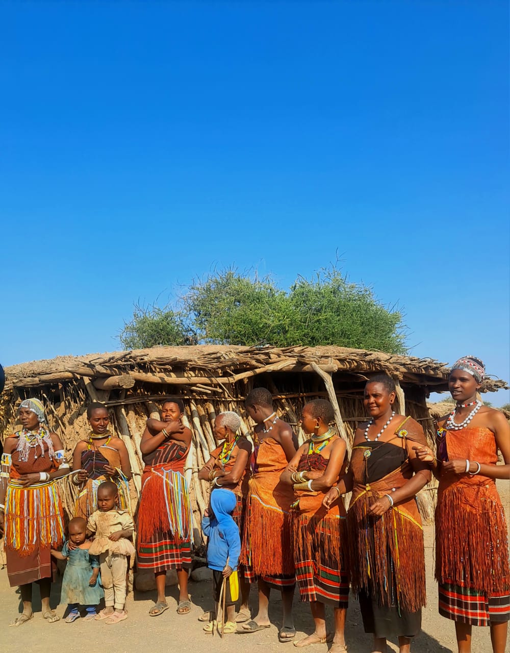 Datoga women and children gathered in beaded dress outside a thatched hut